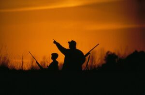 Silhouette of a father and son bird hunting in the orange glow at sunrise.
